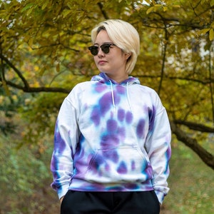 May include: A white tie-dye hoodie with purple and blue accents. The hoodie has a drawstring hood and a kangaroo pocket. The person wearing the hoodie is standing in front of a tree with yellow leaves.