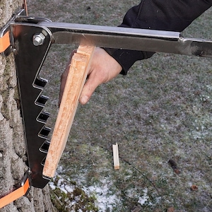 Puede incluir: Una correa de árbol de metal con una palanca y un bloque de madera. La correa está unida a un tronco de árbol con correas naranjas. La palanca se utiliza para apretar la correa.