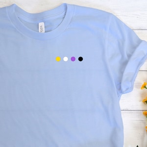 Könnte beinhalten: Hellblaues T-Shirt mit einer kleinen, horizontalen Reihe von vier farbigen Punkten: gelb, weiß, lila und schwarz. Das Shirt ist gefaltet, mit einer blauen Jeans und grauen Turnschuhen im Hintergrund. Die Worte "LIGHT BLUE" sind auf einem Farbfeld aufgedruckt.