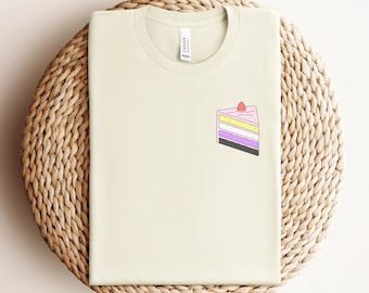 Puede incluir: Una camiseta beige claro con un gráfico de un pastel de capas rosa, amarillo, morado y negro con una fresa en la parte superior. El texto "Natural" está impreso debajo de la imagen.