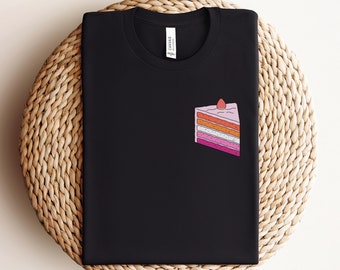 May include: Black t-shirt with a graphic of a slice of layered cake with pink, orange, and white frosting and a red cherry on top.