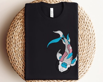 Könnte beinhalten: Schwarzes T-Shirt mit einem blauen, rosa und weißen Koi-Fisch-Design. Der Fisch ist im Stil einer Transgender-Pride-Flaggen-Farbgebung gestaltet.