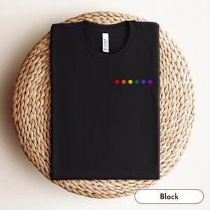May include: Black t-shirt with a crew neck and a small, embroidered rainbow design on the chest. The shirt is folded neatly on a woven, circular mat. The word "Black" is in a white rectangle at the bottom.