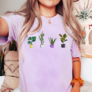 May include: A purple t-shirt with a graphic of five potted plants. The plants include a cactus, a succulent, a monstera, an aloe vera, and a ficus.