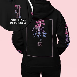 Pode incluir: Moletom preto com um design floral. A parte de trás apresenta um galho de flor de cerejeira rosa e azul dentro de um retângulo rosa, com caracteres japoneses abaixo. O capuz tem texto japonês e as palavras "YOUR NAME IN JAPANESE".