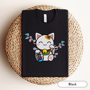 May include: Black t-shirt featuring a cartoon Maneki-Neko, a Japanese lucky cat, holding a skateboard with the words "Nice Try" written on it. The cat is surrounded by pink and blue cherry blossoms.