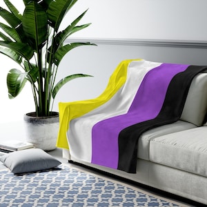 May include: A soft, plush throw blanket featuring the non-binary pride flag colors: yellow, white, purple, and black. The blanket is draped over a light gray couch.