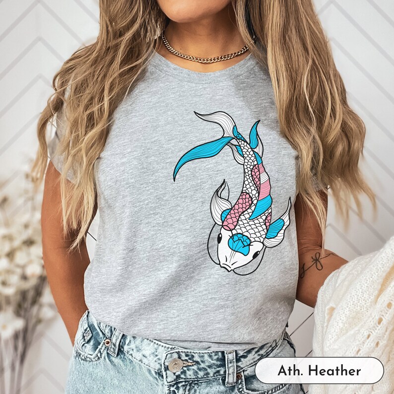 K&ouml;nnte beinhalten: Ein heidefarbenes T-Shirt mit einem blauen, rosa und wei&szlig;en Koi-Fisch-Motiv. Der Text "Ath. Heather" ist in der rechten unteren Ecke des Shirts aufgedruckt.