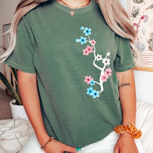 May include: A green t-shirt with a floral design featuring pink, blue, and white flowers. The flowers are arranged in a branch-like pattern on the front of the shirt.