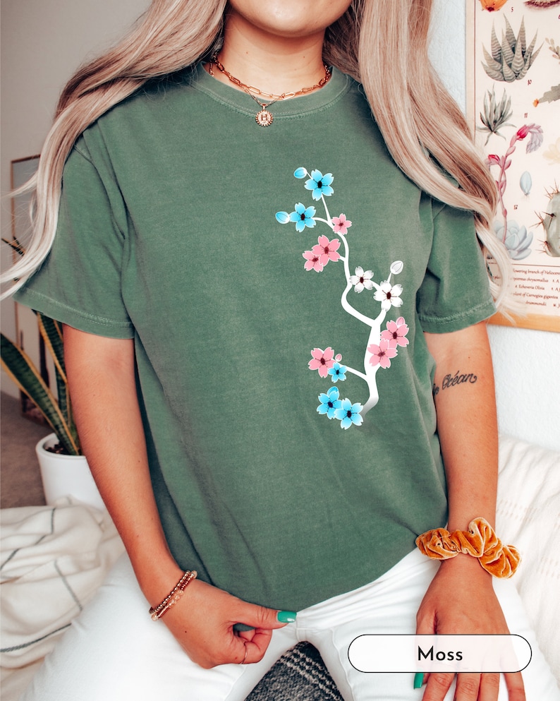 May include: A green t-shirt with a floral design featuring pink, blue, and white flowers. The flowers are arranged in a branch-like pattern on the front of the shirt.