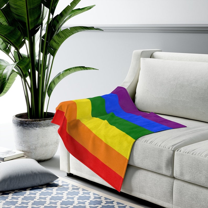 May include: A rainbow-colored throw blanket with stripes of red, orange, yellow, green, blue, and purple. The blanket is draped over a white couch.