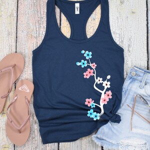 May include: A navy blue tank top with a white floral design. The design features pink, blue, and white flowers on a branch. The tank top is made of a soft, comfortable material and is perfect for everyday wear.