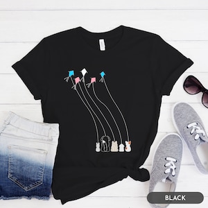 Puede incluir: Camiseta negra con un gráfico de cinco cometas volando en el cielo con cinco animales sosteniendo las cuerdas. Los animales son un conejo, un perro, un gato, un pájaro y un cerdo.