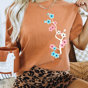 May include: A woman wearing a burnt orange t-shirt with a white floral design and black and brown leopard print shorts. The shirt has a white branch with pink and blue flowers.