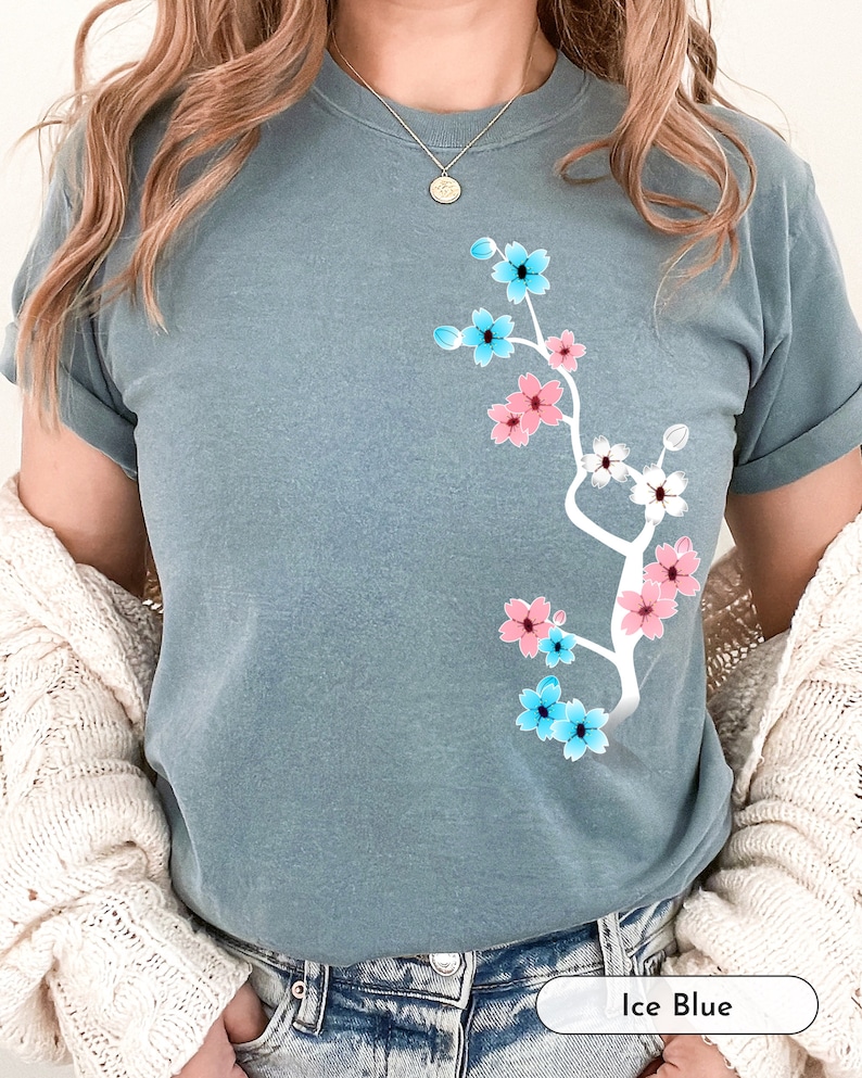 May include: A light blue t-shirt with a white floral design featuring pink and blue flowers. The flowers are arranged in a branch-like pattern. The text "Ice Blue" is visible in the bottom right corner of the image.