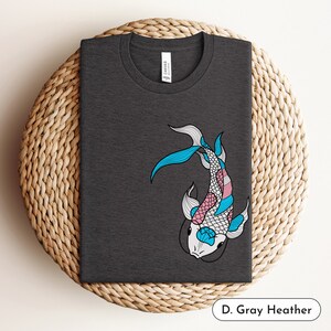 K&ouml;nnte beinhalten: Ein dunkelgraues Heather-T-Shirt mit einem Aufdruck eines blauen, rosa und wei&szlig;en Koi-Fischs. Der Fisch ist im Stil einer Transgender-Pride-Flagge gestaltet.