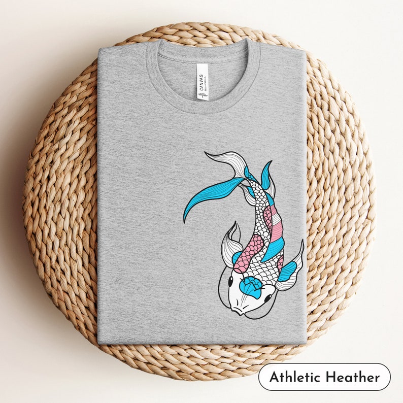 K&ouml;nnte beinhalten: Ein graues T-Shirt mit einem blauen und rosafarbenen Koi-Fisch-Design. Der Fisch ist stilisiert mit einer wei&szlig;en Umrandung und schwarzen Details. Das Shirt ist aus einem weichen, bequemen Material gefertigt und perfekt f&uuml;r den Alltag.