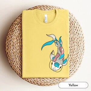 K&ouml;nnte beinhalten: Gelbes T-Shirt mit einem bunten Koi-Fisch-Motiv. Der Fisch ist schwarz umrandet und hat blaue, rosa und wei&szlig;e Schuppen.