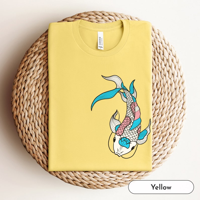 K&ouml;nnte beinhalten: Gelbes T-Shirt mit einem bunten Koi-Fisch-Motiv. Der Fisch ist schwarz umrandet und hat blaue, rosa und wei&szlig;e Schuppen.