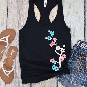 May include: Black tank top with a white branch and pink and blue flowers printed on the front. The text "Black" is printed below the tank top.