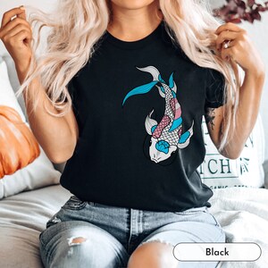 K&ouml;nnte beinhalten: Ein schwarzes T-Shirt mit einem blauen, rosa und wei&szlig;en Koi-Fisch-Motiv. Der Fisch ist stilisiert und hat ein detailliertes Design.