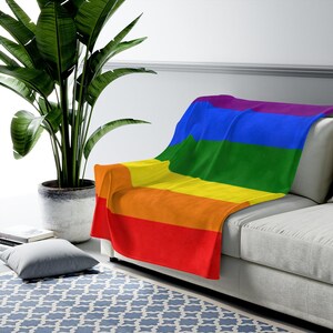 May include: A rainbow-colored fleece throw blanket draped over a light gray couch. The blanket features the colors of the pride flag: red, orange, yellow, green, blue, and purple.
