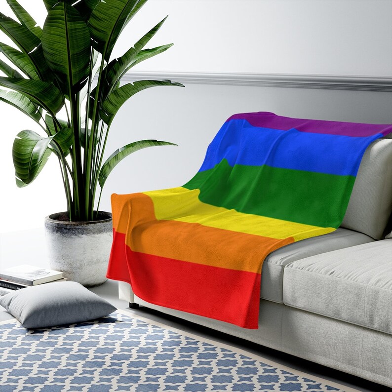 May include: A rainbow-colored fleece throw blanket draped over a light gray couch. The blanket features the colors of the pride flag: red, orange, yellow, green, blue, and purple.