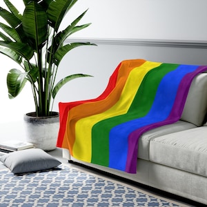 May include: A rainbow-colored fleece throw blanket draped over a light gray couch. The blanket features the colors red, orange, yellow, green, blue, and purple.