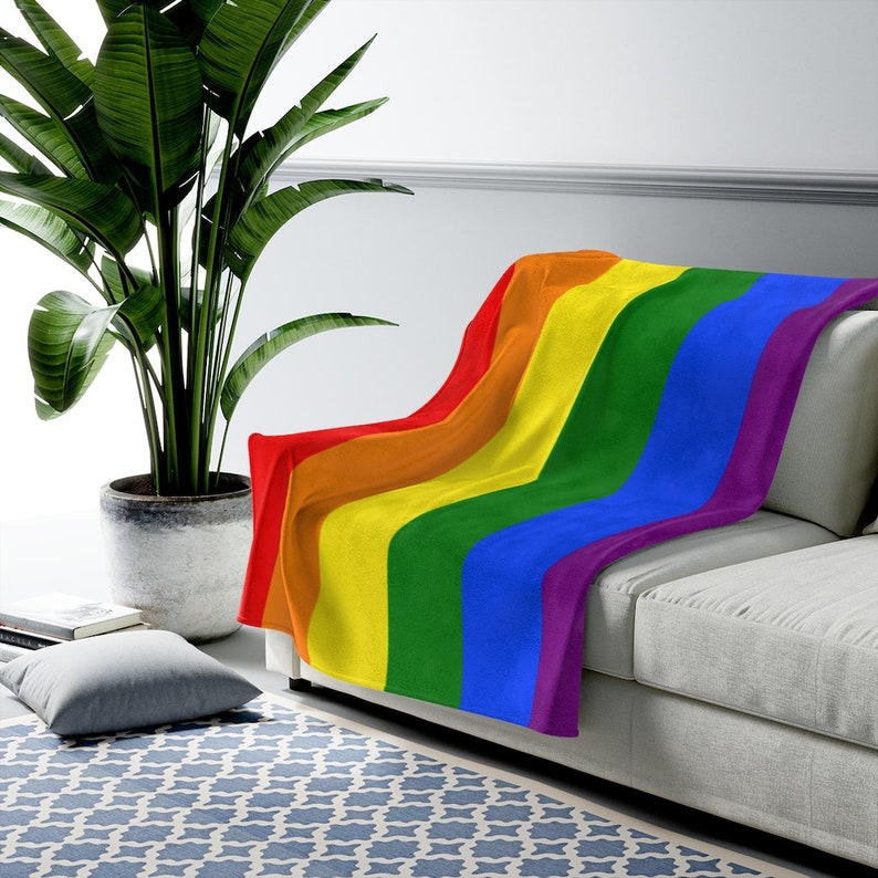 May include: A rainbow-colored fleece throw blanket draped over a light gray couch. The blanket features the colors red, orange, yellow, green, blue, and purple.