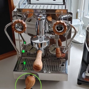 Può includere: Una macchina per espresso in acciaio inossidabile con dettagli in legno. La macchina ha due portafiltri, un manometro e un vassoio di gocciolamento. Un tamper in legno è seduto sul bancone di fronte alla macchina.