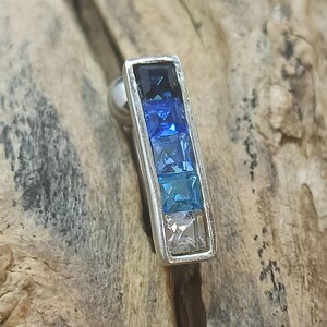 May include: A silver earring with a rectangular design. It features a gradient of square-cut blue gemstones, transitioning from dark blue to light blue and white. The earring has a small, round backing.