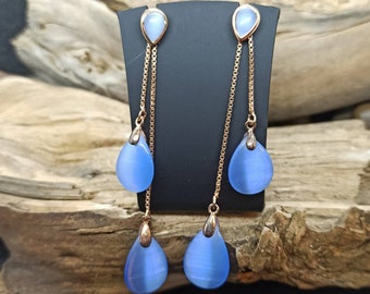 May include: A pair of dangle earrings featuring light blue teardrop-shaped stones. The earrings have a gold-colored chain and accents, with a small, pear-shaped stone at the top. The earrings are displayed on a black stand.