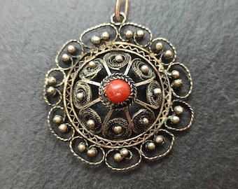Puede incluir: Un colgante de filigrana plateado con una piedra de coral rojo en el centro. El colgante tiene forma de flor con un diseño circular.