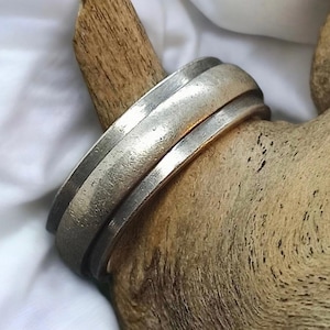 Puede incluir: Un anillo de plata con una banda ancha y una superficie texturizada. El anillo está hecho de dos bandas que están apiladas una encima de la otra.