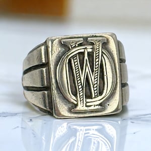 Puede incluir: Un anillo de sello de plata con una cara cuadrada que presenta una letra 'W' grabada dentro de un círculo. La banda del anillo tiene tres líneas elevadas.