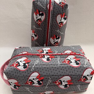 May include: Two gray quilted cosmetic bags with red zippers and handles. The bags feature a pattern of Minnie Mouse in heart-shaped designs. The bags are decorated with red and white polka dot accents.