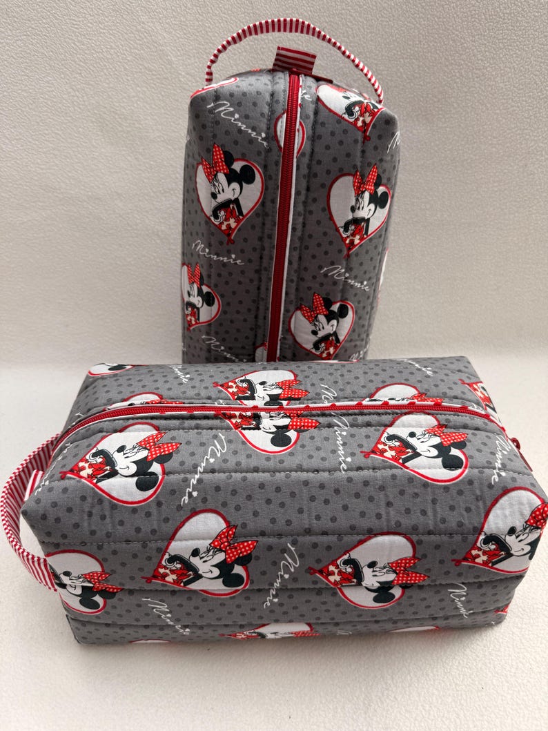 May include: Two gray quilted cosmetic bags with red zippers and handles. The bags feature a pattern of Minnie Mouse in heart-shaped designs. The bags are decorated with red and white polka dot accents.