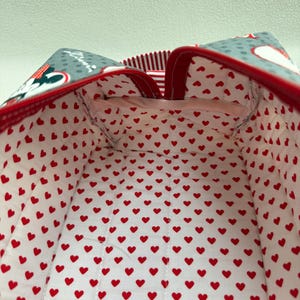 May include: A fabric bag with a red and white heart pattern interior. The exterior features a gray pattern with Minnie Mouse and red trim. The bag's interior is lined with a white fabric covered in small red hearts.