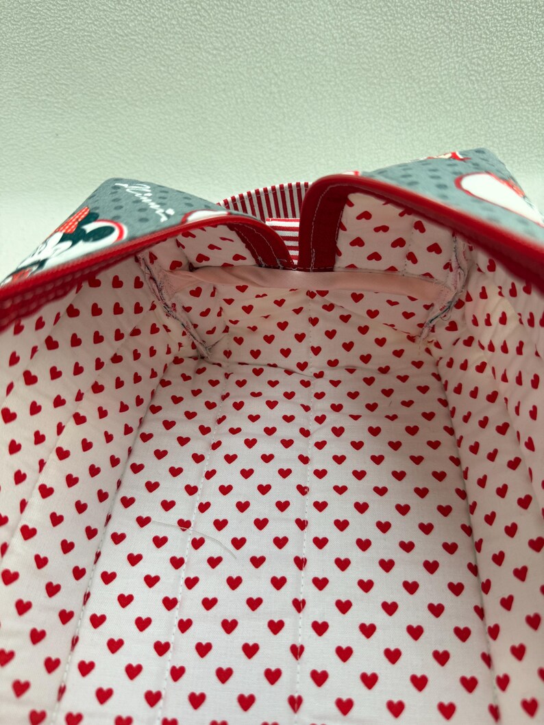 May include: A fabric bag with a red and white heart pattern interior. The exterior features a gray pattern with Minnie Mouse and red trim. The bag's interior is lined with a white fabric covered in small red hearts.