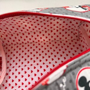 May include: A gray zippered bag with a red trim and a red heart-patterned interior. The exterior features a polka dot pattern and a cartoon character design. The bag is open, revealing the interior lining.