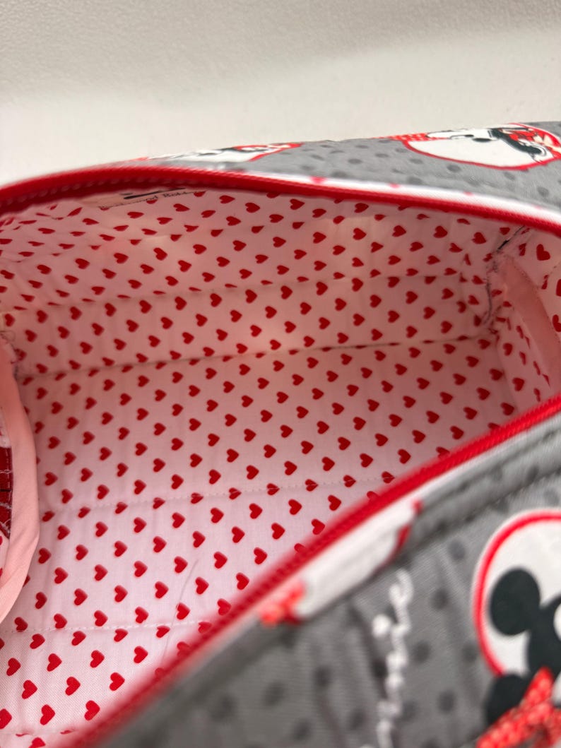 May include: A gray zippered bag with a red trim and a red heart-patterned interior. The exterior features a polka dot pattern and a cartoon character design. The bag is open, revealing the interior lining.