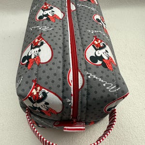 May include: Gray quilted pencil case with a red zipper and a red and white striped handle. The case features a pattern of Minnie Mouse in a heart shape, with the word "Minnie" in cursive.