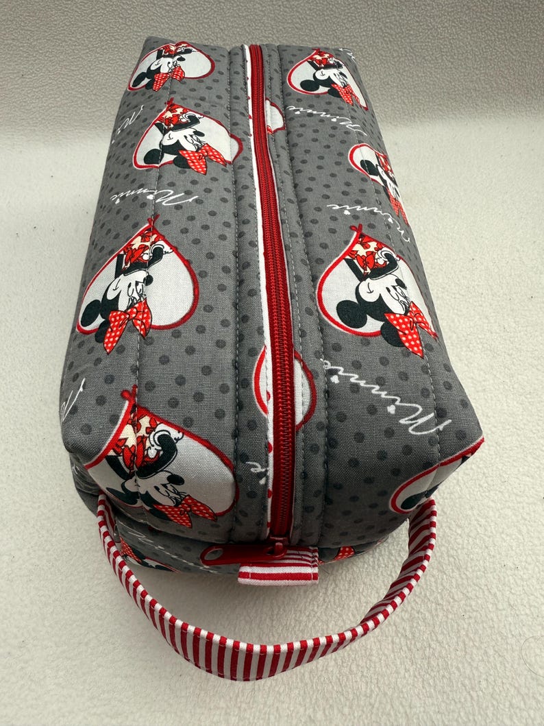 May include: Gray quilted pencil case with a red zipper and a red and white striped handle. The case features a pattern of Minnie Mouse in a heart shape, with the word "Minnie" in cursive.