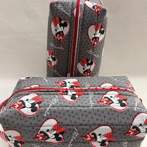 May include: Two gray quilted cosmetic bags featuring a Minnie Mouse heart pattern. The bags have red zippers and handles. The larger bag is rectangular, and the smaller bag is a vertical rectangle. The bags are decorated with a Minnie Mouse design.