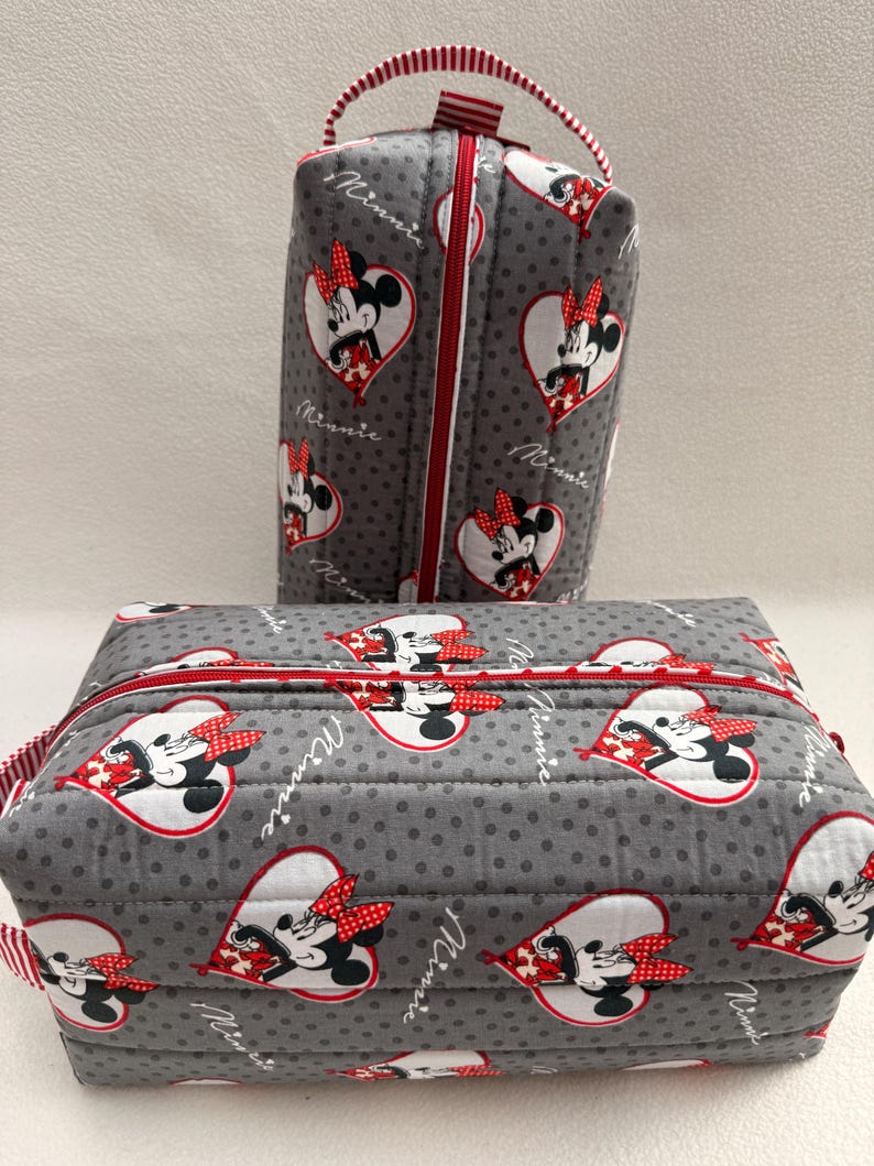 May include: Two gray quilted cosmetic bags featuring a Minnie Mouse heart pattern. The bags have red zippers and handles. The larger bag is rectangular, and the smaller bag is a vertical rectangle. The bags are decorated with a Minnie Mouse design.