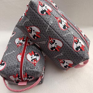 May include: Two gray, quilted cosmetic bags with a Minnie Mouse heart pattern. The bags have red zippers and handles with red and white stripes. The Minnie Mouse design is in a heart shape with the word "Minnie" written on the side.