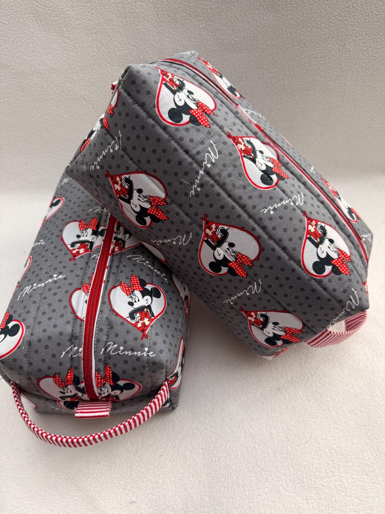 May include: Two gray, quilted cosmetic bags with a Minnie Mouse heart pattern. The bags have red zippers and handles with red and white stripes. The Minnie Mouse design is in a heart shape with the word "Minnie" written on the side.