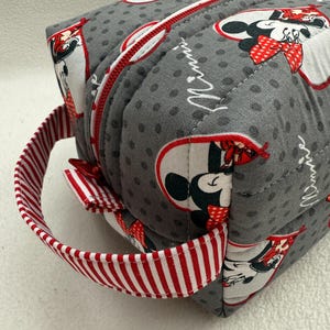 May include: A gray, quilted cosmetic bag featuring a Minnie Mouse pattern. The bag has a red zipper and a red and white striped handle. The bag is rectangular and has a rounded top.
