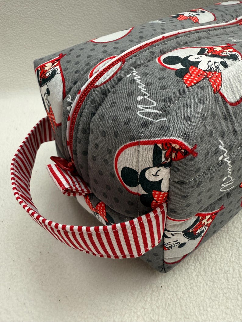 May include: A gray, quilted cosmetic bag featuring a Minnie Mouse pattern. The bag has a red zipper and a red and white striped handle. The bag is rectangular and has a rounded top.