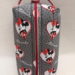 May include: A gray, quilted cosmetic bag featuring a pattern of Minnie Mouse in heart-shaped designs. The bag has a red zipper, a red and white striped handle, and the word "Minnie" in script.